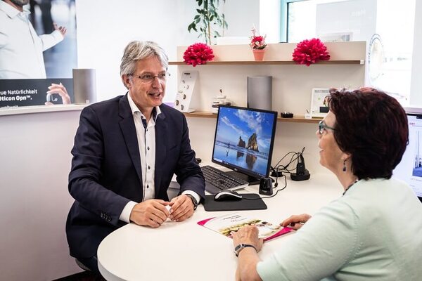 Mitarbeiter im Hörakustik-Fachgeschäft berät Kundin freundlich am Schreibtisch mit Computer.